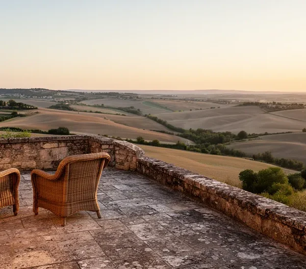Vue panoramique d'une terrasse privée avec deux fauteuils en rotin surplombant les champs de lavande et villages perchés de Provence au coucher du soleil